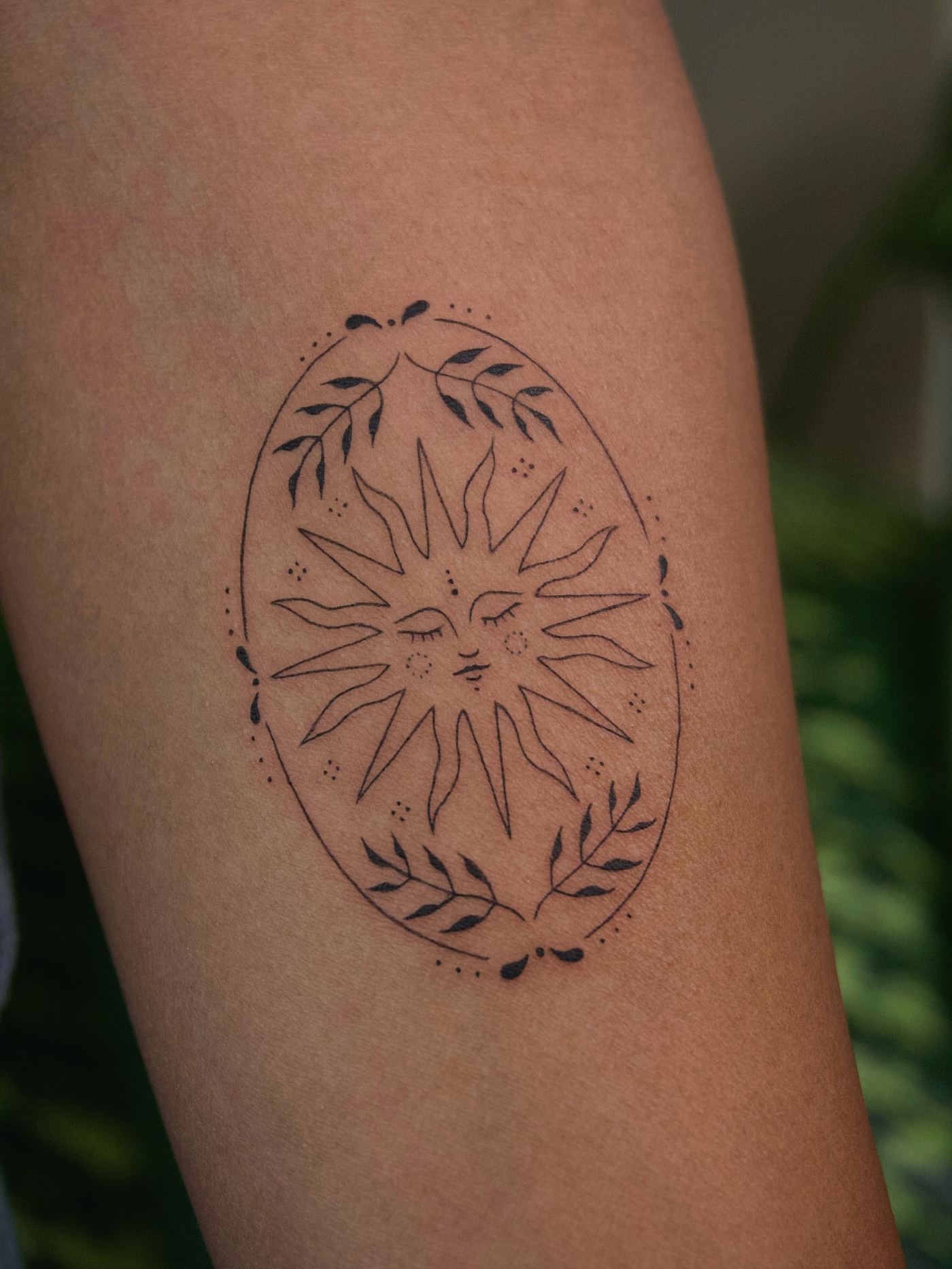 Tatuaje minimalista de un sol con rostro dentro de un óvalo decorado con hojas, brazo