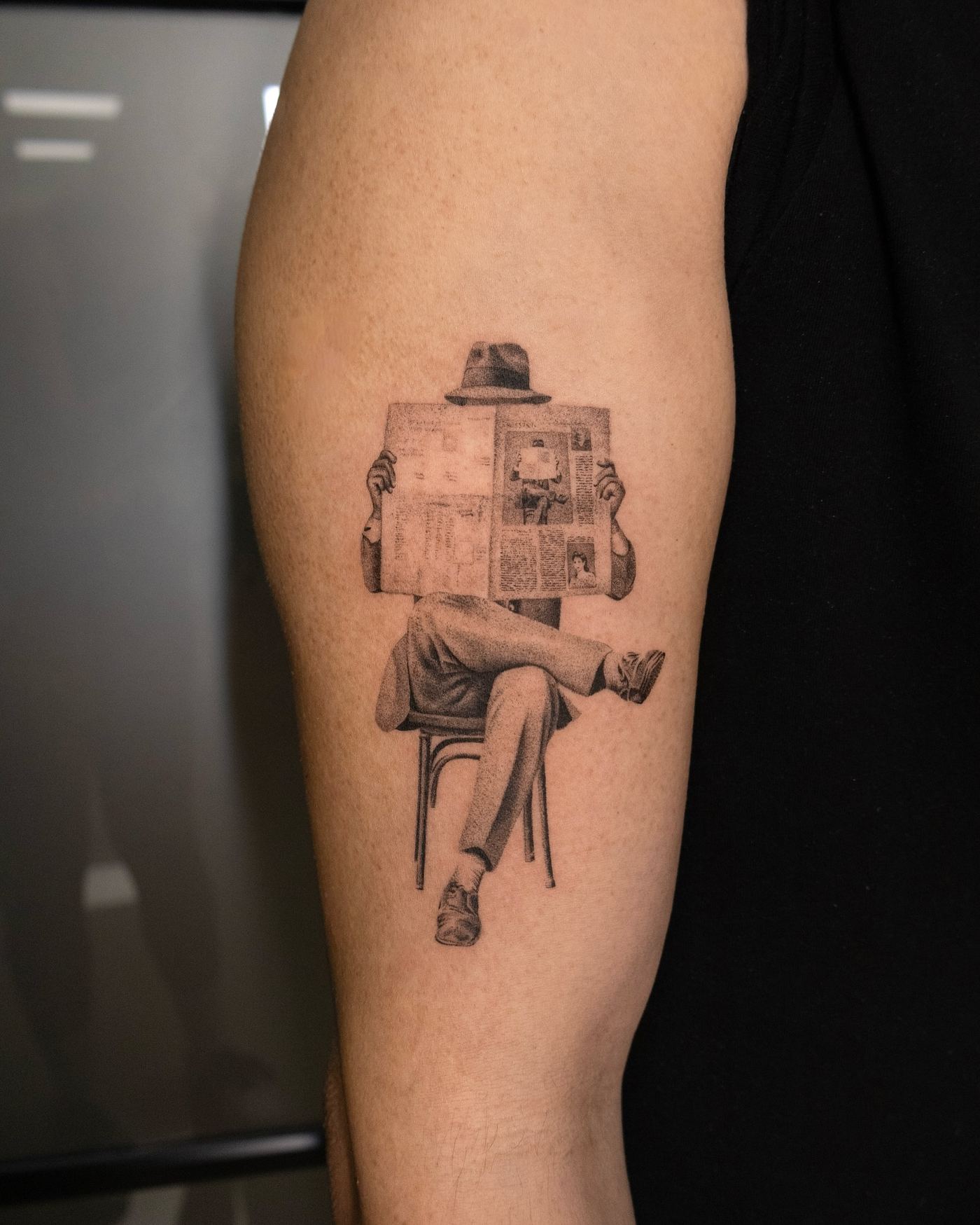 Tatuaje microrrealista de una figura sentada leyendo el periódico, antebrazo